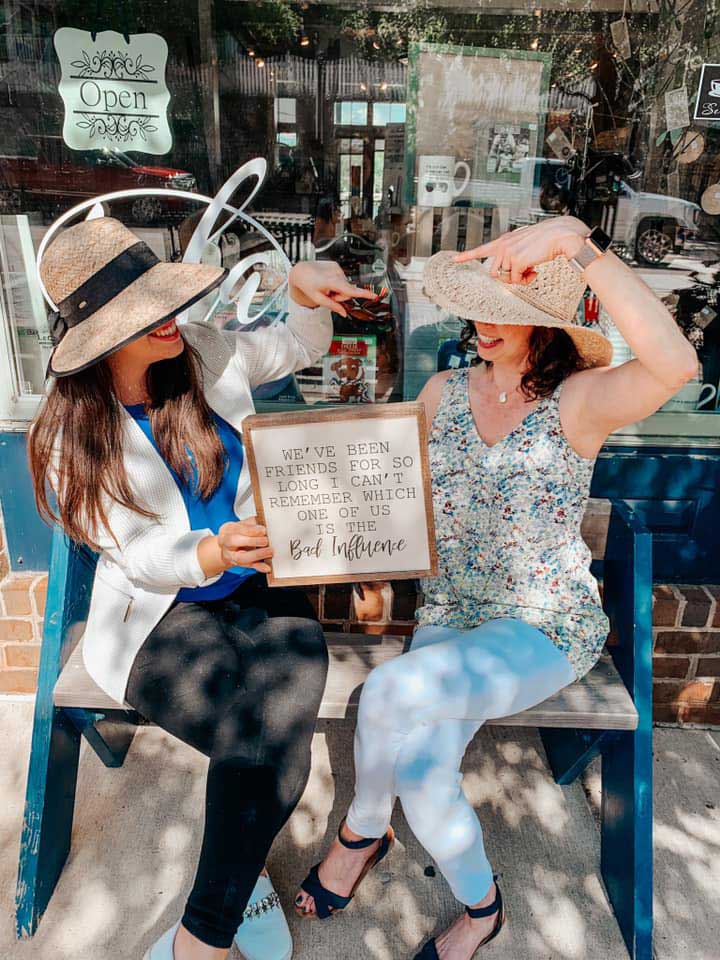 two women wearing large hats sitting on a bench outside of a shop