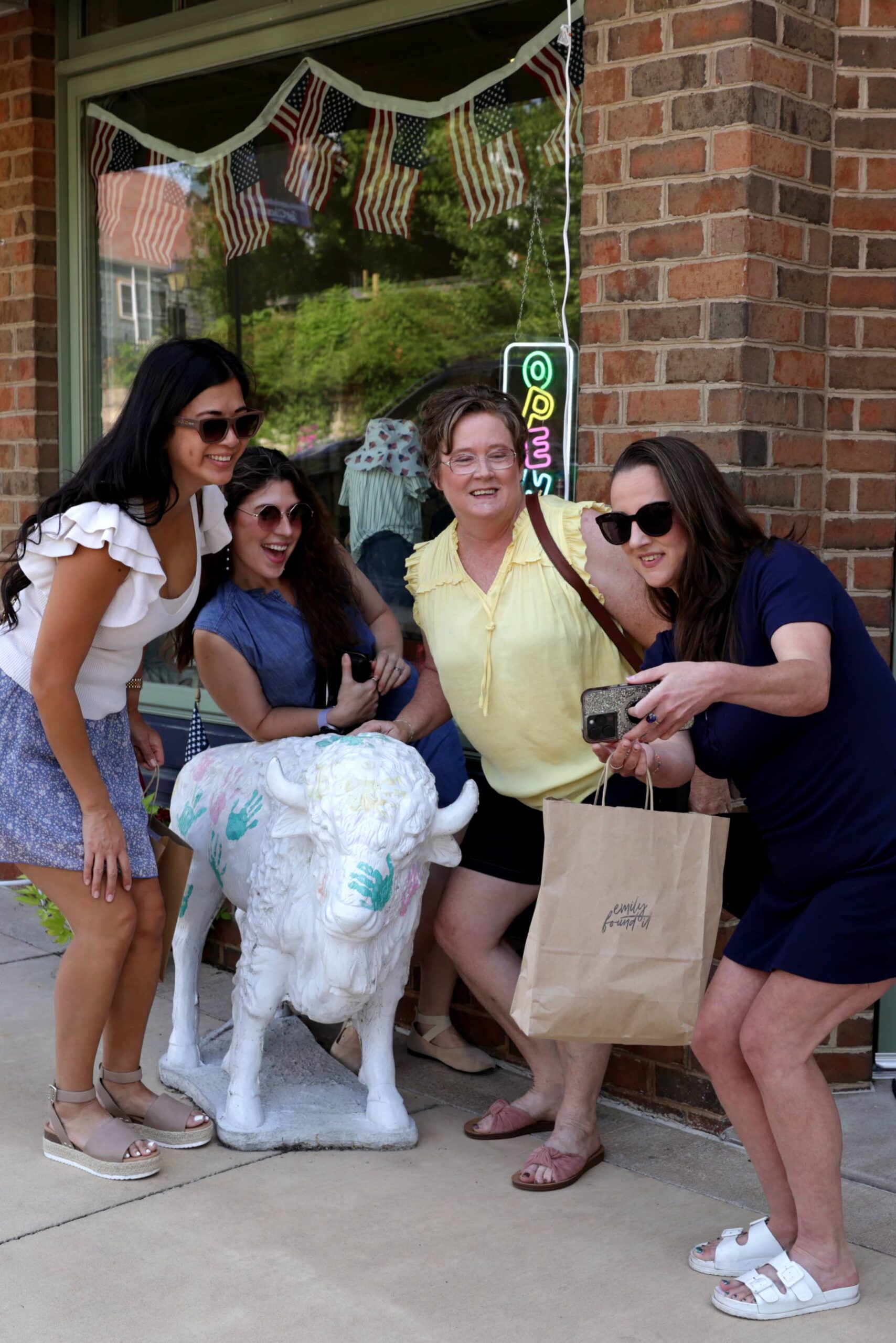 Four women taking a selfie next to a buffalo statue that's been painted white with colorful pastel handprints.